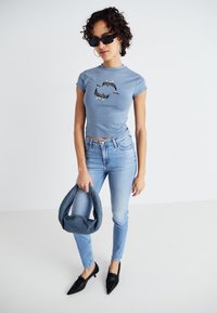T-shirt bleu clair ajusté avec un graphique de dauphin, associé à un jean en denim clair et des chaussures noires à bout pointu. Tenant un sac à main en tissu gris.