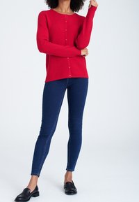 Röd stickad tröja med knappar nedtill, ihop med mörkblå slim-fit jeans och svarta loafers, mot en ljus bakgrund.