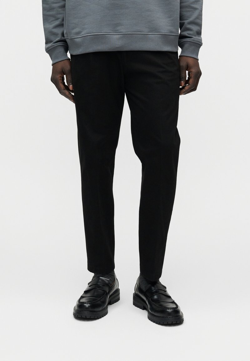 Pantalon noir ajusté en tissu lisse, associé à des mocassins noirs épais. La tenue présente un design simple et moderne.