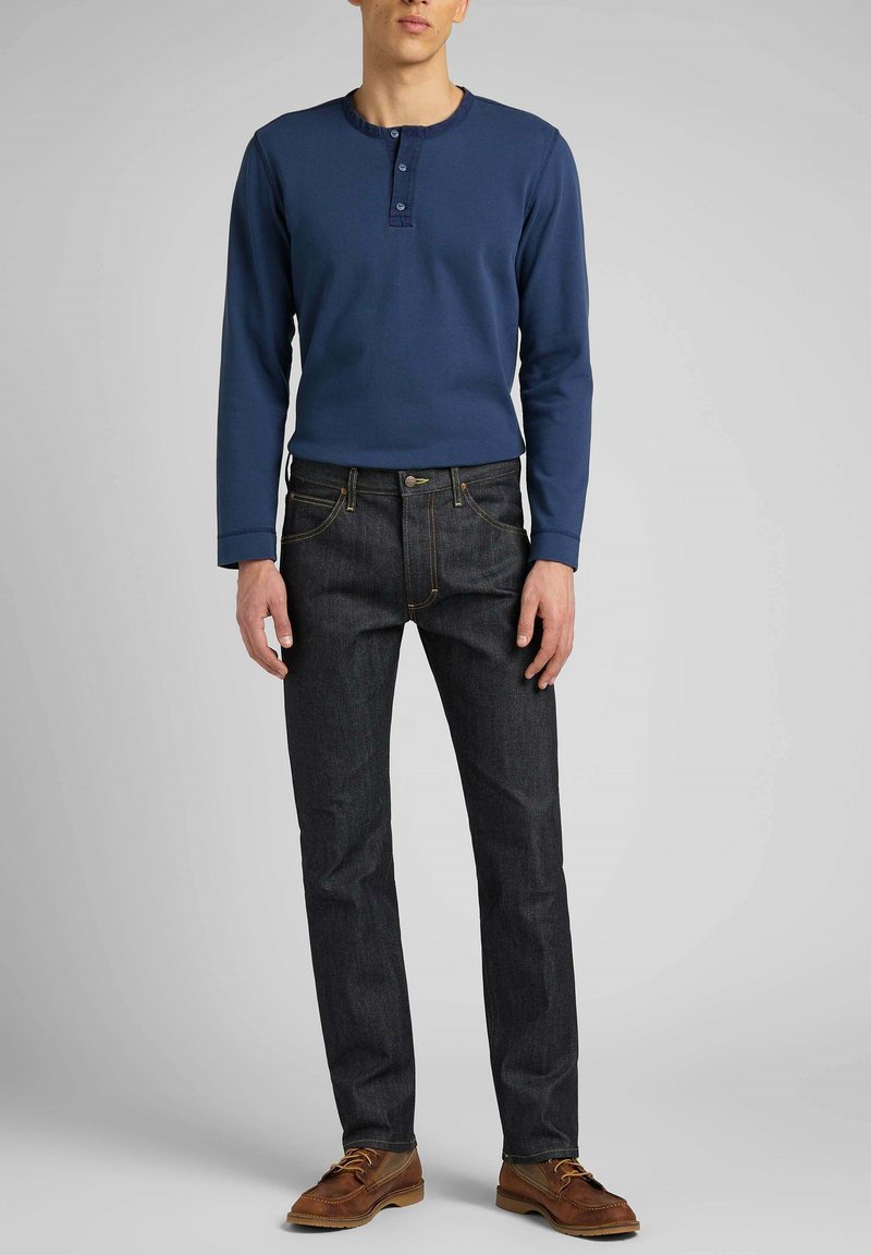 Camisa henley de manga larga en color navy con tres botones, combinada con pantalones vaqueros oscuros y zapatos marrones de cordones. Diseño simple y corte ajustado.
