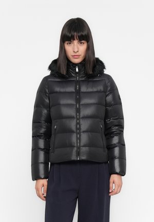 Vrouw in een zwarte pufferjack met een hoge kraag en een met bont afgewerkte capuchon, dichtgeritst, staand tegen een effen witte achtergrond.