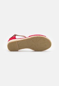 Pohja punaisista espadrille-sandaaleista, joissa on nilkkapanta, varustettuna teksturoidulla kumipohjalla, johon on merkitty "Tommy Hilfiger" -logoja ja -kuvioita.