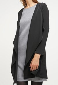Svart, lättviktig, öppen front cardigan med långa ärmar och draperad design, bärs över en grå klänning. Slät textur och minimalistisk stil.