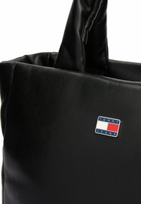 Sort læder skuldertaske med en struktureret overflade, der har to håndtag og et metal logo skilt til Tommy Jeans på forsiden.