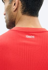 Mies, joka pukeutunut punaiseen paitaan, jossa on teksturoidut pystysuorat raidat ja valkoisella painettu "TÜRKİYE" yläselässä.