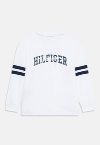 Valkoinen pitkähihaisen polo-paita, jossa on tummansinisiä raitoja hihoissa ja "HILFIGER" painettuna selkään rohkealla fontilla.