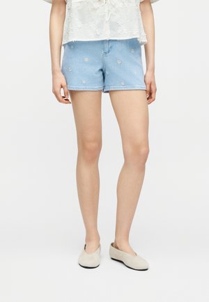 Femme portant un short en jean bleu clair avec de petites broderies de marguerites blanches, un haut fleurie blanc et des chaussures beige à enfiler, debout sur un fond blanc.