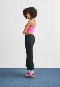 Rosa strikket crop-top, svarte bukser med elastisk midjebånd og rosa joggesko med marineblå striper. Kort, krøllete hår, stående på et blått gulv.