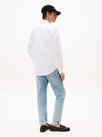 Camisa blanca de manga larga, vaqueros azul claro y mocasines negros. La camisa tiene un cuello abotonado; los vaqueros están enrollados en los puños. Gorra negra.