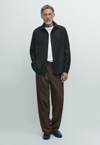 Camicia nera con bottoni, dotata di due tasche sul petto, indossata sopra una t-shirt bianca, abbinata a pantaloni marroni strutturati e scarpe nere.