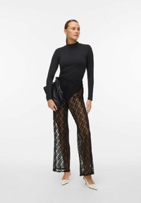 Černý top s dlouhými rukávy v kombinaci s průhlednými krajkovými kalhotami s propracovaným diamantovým vzorem, doplněný černým psaníčkem a bílými podpatky.