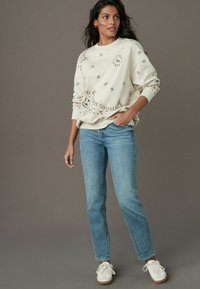 Cream sweatshirt with embroidered details in a sun and swirl pattern, paired with light blue straight-leg jeans and white sneakers.