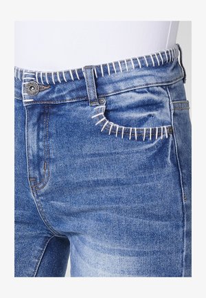 Sinine teksapüksid, millel on unikaalne valge õmblusdetail vöökoha ja taskute ääres, klassikaline viie-taskuline disain ning metallist nööp.