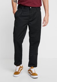 Pantalones negros ajustados de algodón con una textura suave, que cuentan con un cierre de botón y bolsillos laterales, combinados con zapatillas marrones.