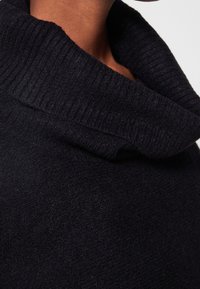 Czarny sweter z dzianiny z szerokim, prążkowanym kołnierzem typu wysoki oraz miękkim, teksturowanym materiałem. Zbliżenie pokazuje szczegóły wzoru dzianiny.