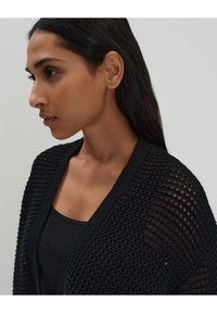 Cardigan noir en tricot avec un design en trame ouverte, présentant un large col en V et des bords texturés. Porté par-dessus un débardeur noir.