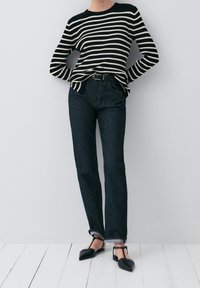 Zwart-wit gestreepte ribbeltrui, donkerblauwe recht-toe-rechtaan jeans, zwarte puntige flats met banden, staand op een witte vloer tegen een lichte muur.