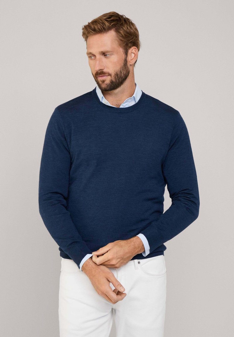 Suéter azul marino, de cuello redondo, con mangas largas, hecho de tejido de punto. Superpuesto sobre una camisa clara de color azul con cuello, combinado con pantalones blancos.