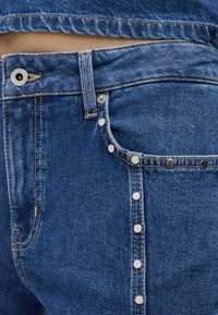 Denim jeans in een donkerblauwe kleur, met zilveren drukknoopsluitingen aan de zijkant en zichtbare stiksels. Zachte, gestructureerde stof.