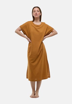 Femme debout pieds nus, mains sur les hanches, portant une robe marron ample mi-longue à manches courtes sur un fond uni.