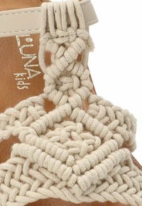 Sandalia tejida en color beige con un diseño trenzado sobre una suela marrón. Incluye una correa con detalles de nudos decorativos.