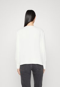 Weiße Bluse mit langen Ärmeln, geradem Saum und geknöpften Manschetten, kombiniert mit dunkelgrauen Jeans. Glattes, ungemustertes Material.