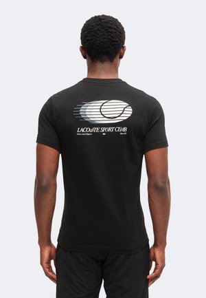 Mann trägt schwarzes T-Shirt mit weißem Tennisball-Motiv und dem Aufdruck „Lacoste Sport Club“ auf dem Rücken, steht mit dem Rücken zur Kamera.