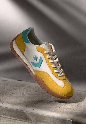 Bílá teniska se žlutými semišovými akcenty, tyrkysovým logem ve tvaru hvězdy, gumovou podrážkou a bílými tkaničkami, zobrazena na betonovém povrchu s jemnými stíny.