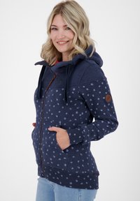 Sweatshirt à capuche bleu marine avec motif à pois, fermeture éclair à l'avant, deux poches et un écusson logo marron sur la manche gauche. Tissu doux, coupe décontractée.