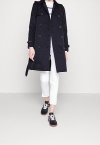 Svart trenchcoat med dubbelknäppt fram, axelpartier och kurvad nederkant, i kombination med vita cropped byxor och svarta sneakers.