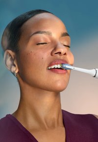 Spazzolino elettrico con manico bianco e testina blu, tenuto vicino a una bocca aperta. Texture della pelle liscia e trucco sottile sulla persona.