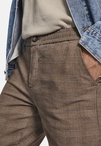 Braun karierte Hosen aus strukturiertem Stoff, mit maßgeschneiderter Passform, Fronttaschen und Knopfverschluss. Getragen mit einem hellen Hemd und einer Jeansjacke.