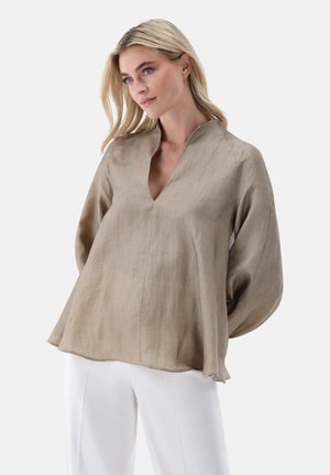 Blonde Frau, die eine lockere beige V-Ausschnitt-Bluse mit langen Ärmeln und weiße Hose trägt, steht mit den Händen hinter dem Rücken vor weißem Hintergrund.