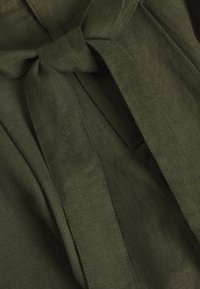 Olivgrön tyg med en strukturerad yta, med detaljer i form av fluga och lösa draperingar. Materialet verkar vara lätt och ventilerande.