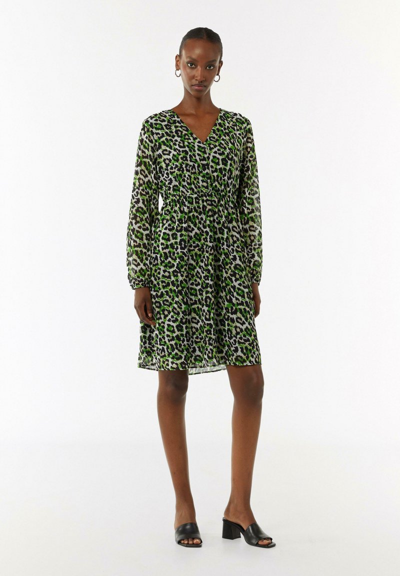 Grünes Leopardenmuster-Kleid mit langen transparenten Ärmeln, V-Ausschnitt, gerafftem Taillenbereich und knielangem Saum, kombiniert mit schwarzen Absatzsandalen.