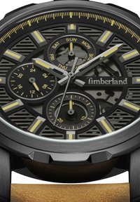 Montre Timberland avec un cadran noir texturé, des accents jaunes et deux sous-cadrans. Elle possède une lunette grise et un bracelet en cuir marron.