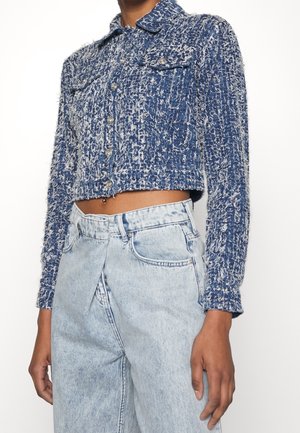 Mujer vistiendo una chaqueta corta texturizada azul y blanca con botones plateados y jeans de talle alto de lavado claro sobre un fondo liso.