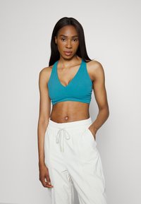 Sutiã desportivo em teal com decote em V, alças largas e design sem costuras. Usado com calças soltas de cor clara, com cordão.