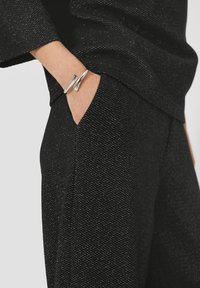 Main dans une poche de pantalon noir texturé, portant un bracelet en argent abstrait, faisant partie d'une tenue assortie en noir texturé.