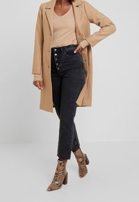 Cappotto lungo color cammello su un top beige, abbinato a jeans neri a vita alta con dettagli sui bottoni e stivaletti alla caviglia con stampa serpente.