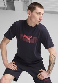 Jūras zila kokvilnas T-kreklis ar lielu sarkanu "PUMA" logo, kas izceļ atkārtotu tekstu; modelis valkā koordinējošas šortus, atklājot tetovējumu uz rokas.
