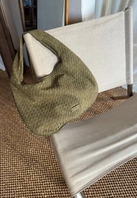 Sac à épaule tissé vert olive reposant sur une chaise en tissu léger. Présente une finition lisse et un subtil détail de logo à l'avant.