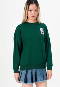 Sweatshirt vert avec un col rond et des épaules tombantes, arborant un logo "G" rose. Associé à une jupe en denim plissée avec des bords effilochés.