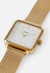 Montre carrée dorée avec un cadran blanc, comportant des index des heures dorés, et un bracelet en maille. La couronne est sur le côté droit. Nom de la marque : CLUSE.