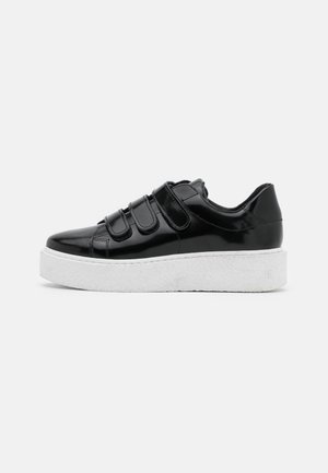 Black patent leather sneaker with three Velcro straps and thick white platform sole on plain background.