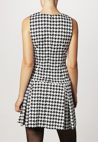 Nainen, joka käyttää mustavalkoista houndstooth-kuvioista mekkoa ilman hihoja, näkyy takaa mustien sukkahousujen kanssa.