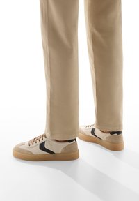 Le sneakers presentano una tomaia in pelle e suede beige, dettagli neri e una suola in gomma color caramello. I lacci sono marroni, con una punta rotonda e un design a profilo basso.
