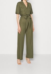 Olivgrön jumpsuit med korta ärmar, knäppt skjortkrage, sidfickor, bälte i midjan och byxdessign med vida ben. Buren med nude klackar.
