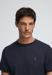 T-shirt in cotone blu navy con scollo rotondo, caratterizzata da un piccolo logo rosso ricamato di un golfista sul petto a sinistra. Texture liscia.