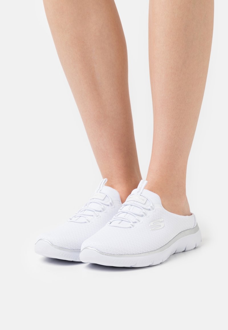 Baskets blanches sans fermeture avec lacets portées pieds nus, avec une semelle rembourrée et un logo latéral sur fond blanc.